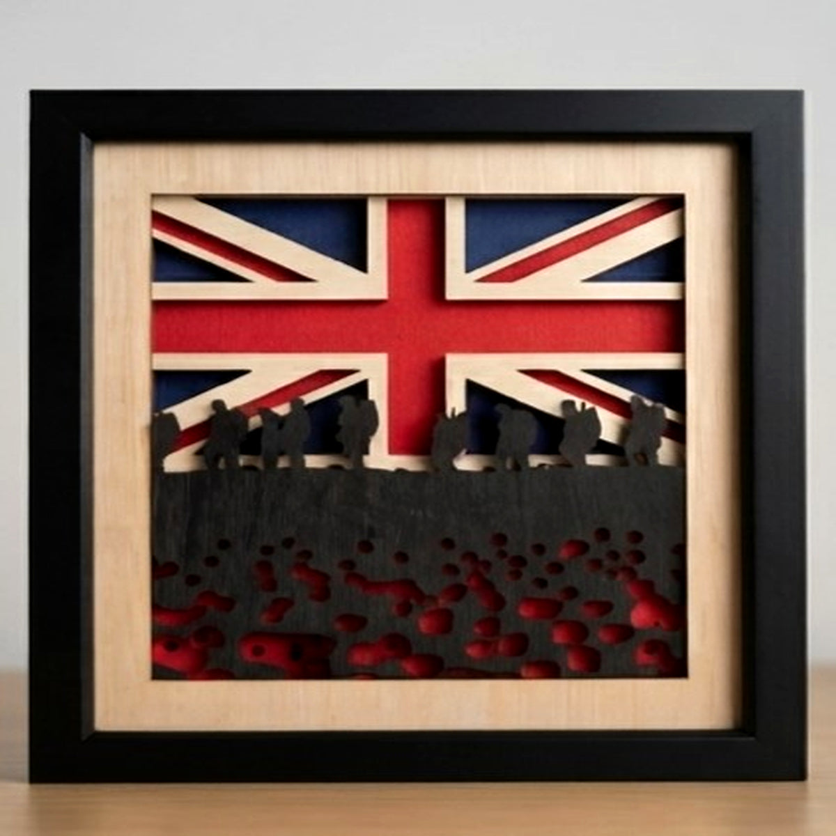 A square framed wooden wall art piece featuring a layered Union Jack flag design, dark silhouette figures across the middle, and red cutout details near the bottom, displayed against a light gray wall.