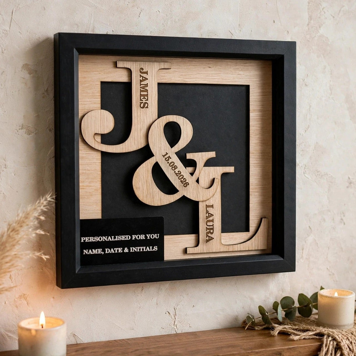 A personalized wooden monogram shadow box framed in black, mounted on a textured beige wall above a wooden surface with lit candles and dried grass decor. The monogram features large engraved letters J, A, and G with names James, Amanda, and Gavin, plus a black plaque area for personalization.