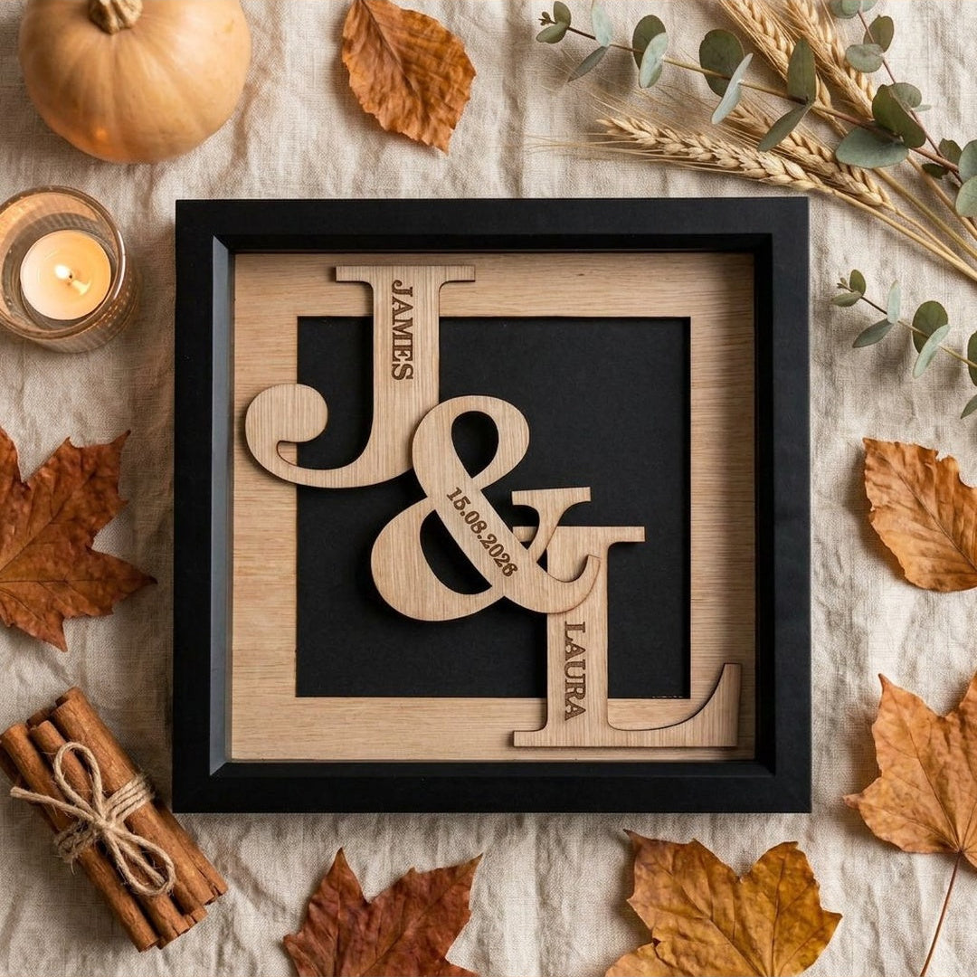 A framed personalized wooden monogram wall art piece with the names James, Sarah, and Liam integrated into large decorative letters, styled in a flat lay with autumn leaves, a candle, cinnamon sticks, and a small pumpkin on a neutral fabric background.
