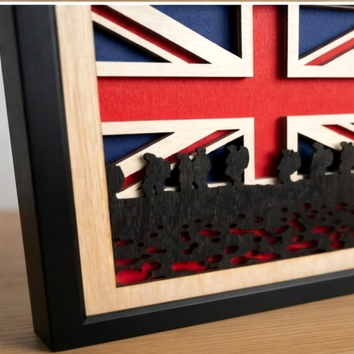 A close-up of a framed layered wooden wall art piece featuring a Union Jack flag design with silhouette figures along the bottom, shown on a light wood surface.